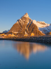 Vertical view of Reine and Hamnoy fishing villages at sunrise, pink glowing mountain peaks and traditional rorbu cabins. Crystal reflections on calm fjord water, Lofoten Islands, Norway.