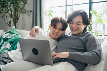 Relaxed Asian gay couple sitting on living room sofa and watching laptop together. A warm domestic lifestyle moment showing digital connection, comfort, and modern home entertainment for two men