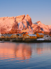 Vertical view of Reine and Hamnoy fishing villages at sunrise, pink glowing mountain peaks and traditional rorbu cabins. Crystal reflections on calm fjord water, Lofoten Islands, Norway.