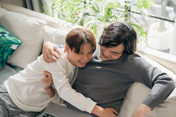 Relaxed Asian gay couple sitting on living room sofa and watching laptop together. A warm domestic lifestyle moment showing digital connection, comfort, and modern home entertainment for two men