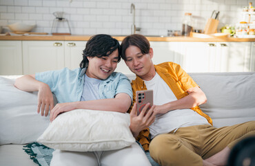Cheerful Asian gay couple taking a selfie with smartphone while leaning together on sofa at home.
