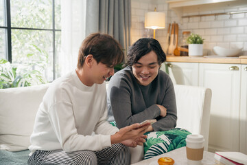 Happy Asian gay couple sitting on living room sofa, smiling while sharing a smartphone. A warm domestic lifestyle moment showing connection and modern comfort at home for two men using technology