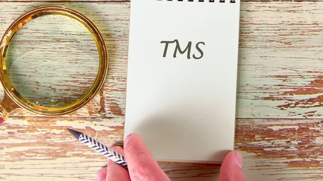 first someone sketches letters on paper, then traces the letters TMS on a notepad with a pen. A magnifying glass sits nearby on the antique table