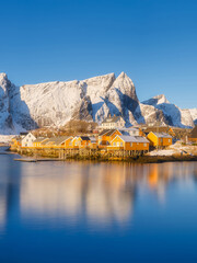 Vertical view of Reine and Hamnoy fishing villages in bright early morning light, traditional rorbu cabins and snowy mountain peaks. Crystal reflections on calm fjord water, Lofoten Islands, Norway. 
