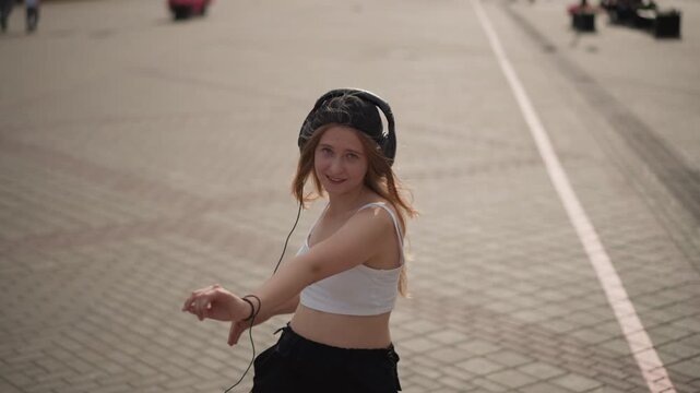 young white woman dancing on street, sunlit pedestrian plaza, wearing crop top and headphones, twirling and smiling, carefree movement, long hair flowing, headphones cable visible, casual black