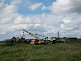 Fototapeta premium Old industrial crane in a junkyard with overgrown grass and various abandoned machinery under a cloudy sky, showing decay.