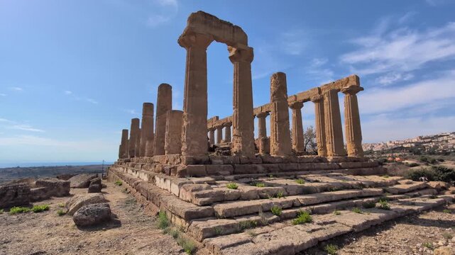 Slow leftward pan across the Temple of Juno in the Valley of the Temples, showing Doric columns, stepped foundations, and the open landscape under clear Mediterranean skies.