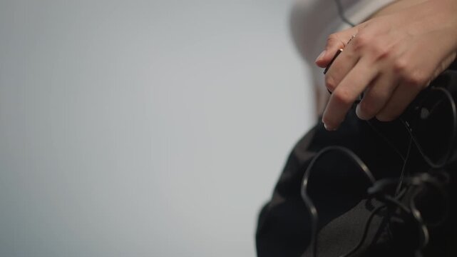 young caucasian woman adjusting wired player, sunlit closeup of hands untangling earphone cord and tapping playlist on blue device, wearing white crop top and black joggers, subtle hip movement