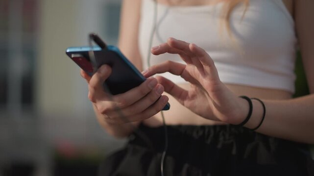 young female street adjusting phone music player, earphones plugged into device, scrolling playlist between rehearsals, cropped white top and black pants, sunlit urban background, closeup on hands