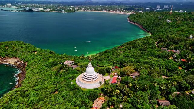 Aerial drone shot of the white Japanese Peace Pagoda rising above lush Rumassala forest and turquoise Galle Bay on Sri Lanka&rsquo;s south coast, with distant seaside town along the horizon.