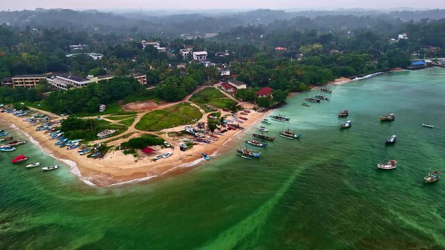 Drone aerial of Weligama fishing village on Sri Lanka&rsquo;s south coast, with colorful boats lining a sandy peninsula and floating in a shallow turquoise bay.