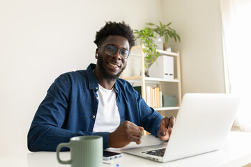 Happy African American man working at home office using laptop looking at camera smiling.