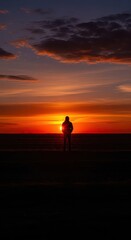 Fototapeta premium Solitary figure stands silhouetted against a brilliant orange sunset over open terrain
