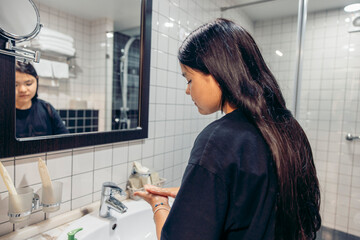 Young Asian woman with long black hair washing her hands at a bathroom sink. The scene emphasizes personal care and hygiene in a modern setting.