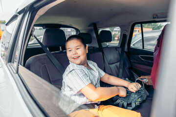 A young Asian boy sits in the back seat of a car, smiling and holding a toy. The interior is modern and spacious, indicating a family trip.