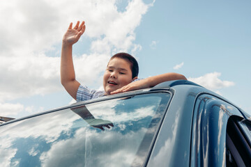 A young Asian boy waves from the open window of a car during a road trip. The sky is blue with fluffy clouds, creating a cheerful atmosphere.