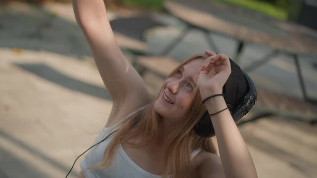 young woman dancing outdoors with headphones, eyes closed and smiling in sunlit park wearing beanie and white tank top, gentle arm movements, relaxed sway near picnic bench, mellow summer vibe,