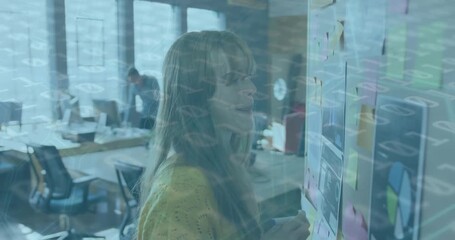 A young woman is writing on a mood board full of graphs and sticky notes in a modern office - Powered by Adobe