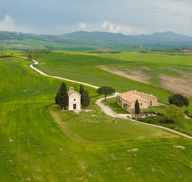Aerial view of a rustic chapel and farmhouse nestled in the vibrant, rolling green hills of the Tuscan countryside, with winding white roads under a dramatic sky, Pienza, Tuscany, Italy.