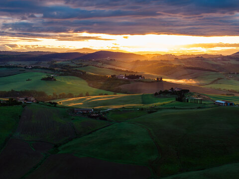 Aerial view of sunbeams piercing through the clouds over the rolling green hills and farms, casting long shadows across the landscape, Crete Senesi, Italy.