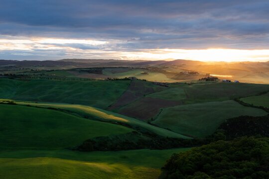 Aerial view of rolling green hills bathed in the soft golden light of dawn under a vast sky, Crete Senesi, Italy.