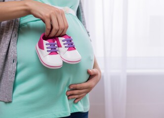 A pregnant woman in a teal shirt holding baby shoes against her belly
