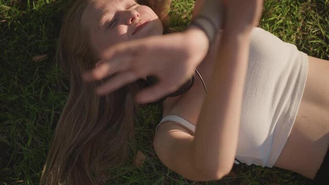 young woman lying on grass listening to music with headphones around neck, sunlight warming face, eyes closed smiling, slow hand gestures, long hair spread on lawn, crop top, barefoot summer vibe,