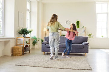 Mother and daughter dancing at home together. They play and spin by sofa in living room, laughing and holding hands. Parent and child girl having fun indoors. Celebrates joy and family bonding.