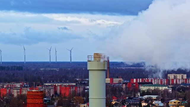 Drone orbiting around tall industrial smokestack with vapor and wind turbines