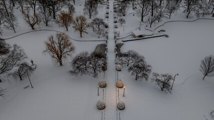 Aerial view of a snow-laden Boston Public Garden, where a blanket of white softens the park's pathways and trees, contrasting with the warm glow of streetlights, Boston, Massachusetts, United States.