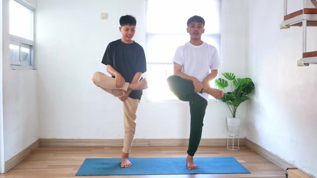 Two teenage brothers performing various standing stretches and balance exercises on blue yoga mat at home.