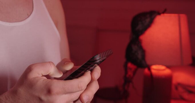 a person typing on a smartphone in a moody red-lit bedroom with lingerie on a lamp, symbolizing discreet online flirting and private communication.