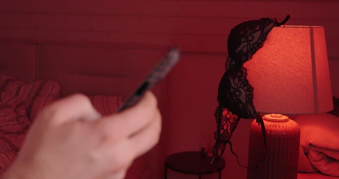 A person uses a smartphone in a red-lit bedroom while lingerie hangs on a lamp, suggesting private messaging, online flirting, and discreet romantic communication.