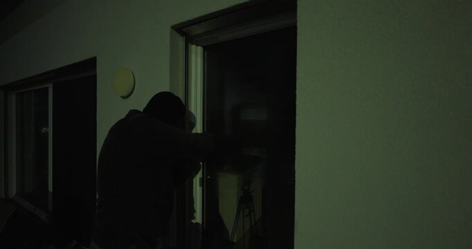 Close-up of a hooded intruder at night attempting to open a patio sliding door from outside, creating a tense home-security and burglary concept.