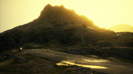 A winding road leads through rugged terrain as the sun sets behind a majestic mountain. The warm...