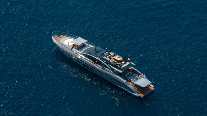 Aerial shot of a sleek, modern yacht cruising across deep blue water. The luxury vessel features a metallic silver hull and multiple wooden decks with lounge areas. Concept of luxury boat holidays. 