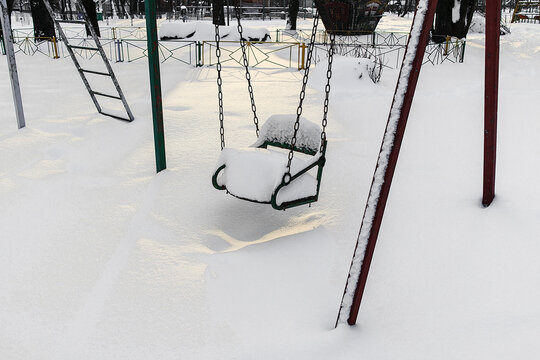 Snow-covered playground swing standing still in winter, showing cold weather, silence, and absence of children in an outdoor park.