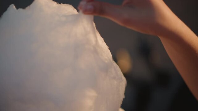 closeup cotton candy hand pulling fluff, soft illuminated fair stall scene, fingers teasing sticky sugar cloud, bokeh lights, warm evening glow, delicate texture, satisfying bite, carnival atmosphere,