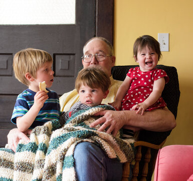Three happy young children sitting under a blanket on their grandfather's lap in an armchair