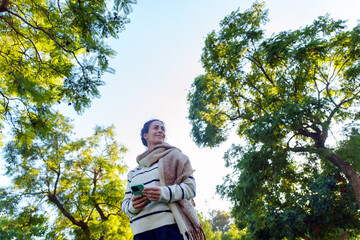 Woman smiling, looking up while holding a phone in a park, enjoying nature and digital connection
