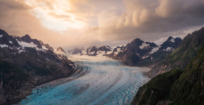 Aerial view of a radiant glacial river flowing through rugged mountains under a sky of soft light and looming clouds, Cordova, Alaska, United States.