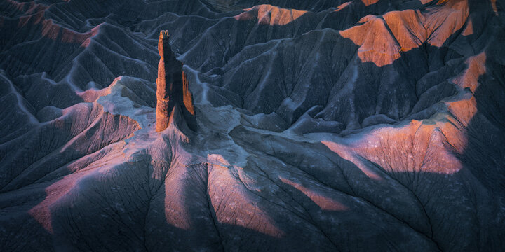 Aerial view of a solitary spire bathed in the warm glow of sunset amidst the rugged, textured landscape, Hanksville, Utah, United States.