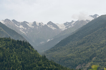 Majestic mountain range with dense forested slopes and distant snow capped peaks beneath a misty cloudy sky and valleys stretch into distance.