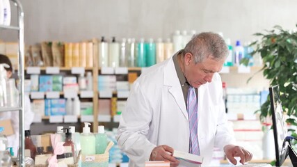 Senior male pharmacist uses computer at workplace. Employee reads regulations and laws, makes report, reads information about drugs. Girl colleague work in background