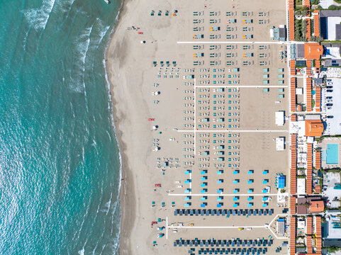 Aerial view of turquoise waves gently kiss the sandy shore, meeting neat rows of beach umbrellas and buildings, Forte dei Marmi, Tuscany, Italy.