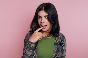 Young person stands against pink wall, thinking with hand on mouth in casual clothing in indoor setting during daytime