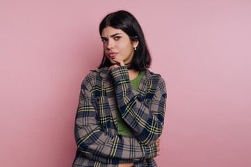Fototapeta premium Young person standing against pink background showing thoughtful expression while posing with arms crossed and hand on chin