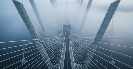 Cinematic drone view looks down on geometric suspension bridge cables and supports crossing a foggy river at cool grey dawn