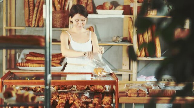 Young female shopper selects and places fresh croissants in plastic bag at a bakery. High quality 4k footage