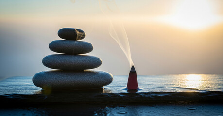 Zen stacked polished river stones and smoking incense rest on dark slate backlit by a golden sunset